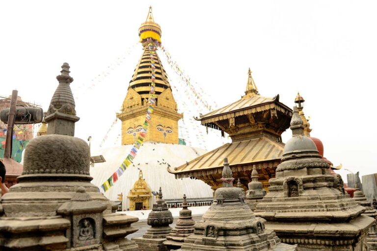 Stone stupas and pagoda structures in front of Swayambhunath Stupa, also known as the Monkey Temple, with prayer flags and the stupa’s eyes visible in the background.