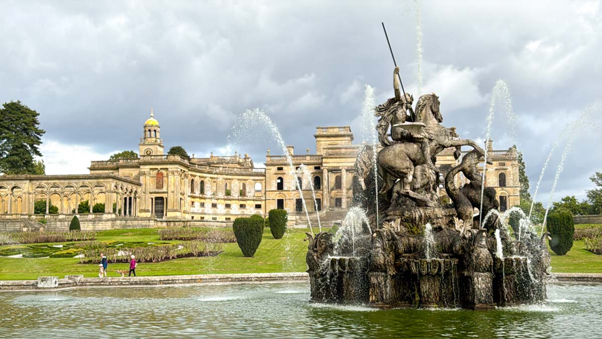 A large ornate fountain with statues is in front of a grand historic building, with trimmed hedges and a cloudy sky in the background.