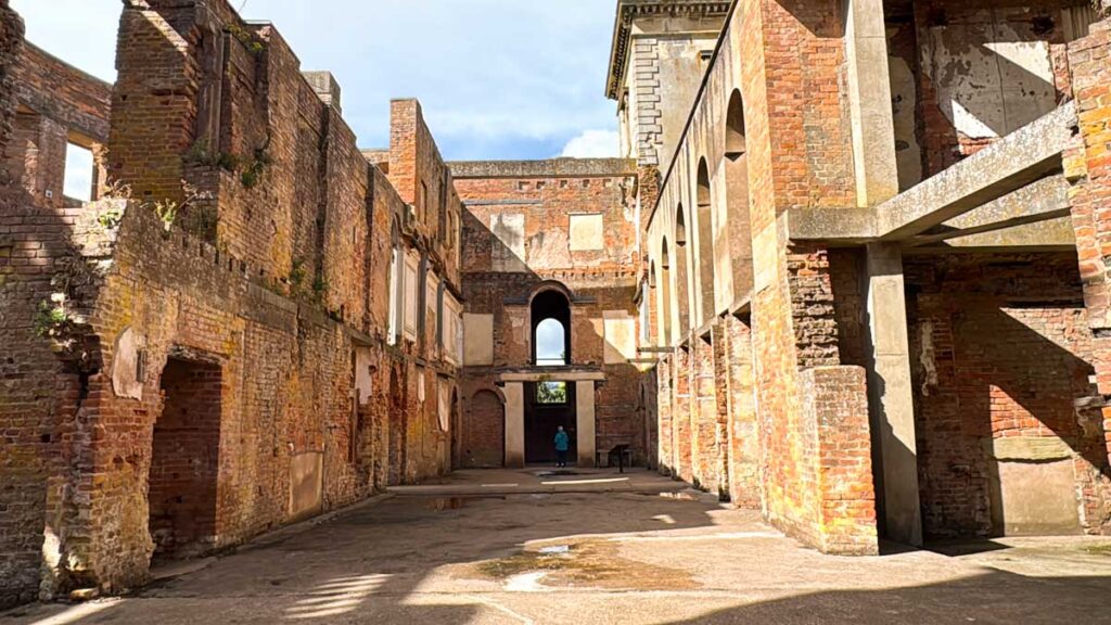 Interior view of a brick and stone building ruin with missing roof, exposed walls, and arched windows; a person stands in the distance at the far end.