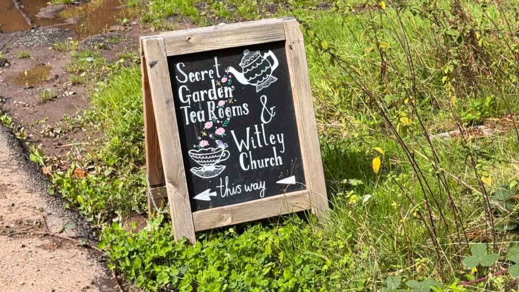 A wooden sign on grass reads "Secret Garden Tea Rooms & Witley Church this way" with illustrations of a teacup, cake, and flowers.