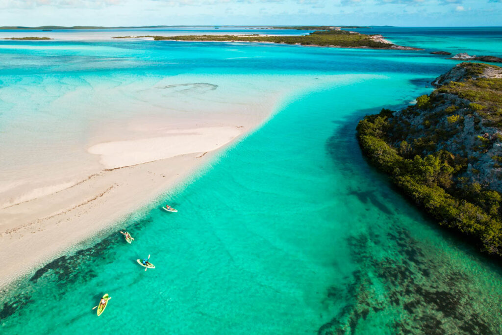 Aerial view of people kayaking in clear turquoise water near a sandy shore and lush green island coastline under a partly cloudy sky.