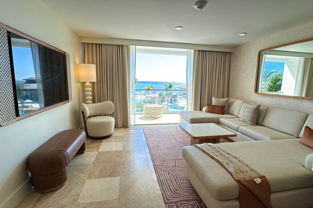 Modern hotel suite with sectional sofa, armchair, TV, and large window opening to a balcony with a view of palm trees and the ocean.