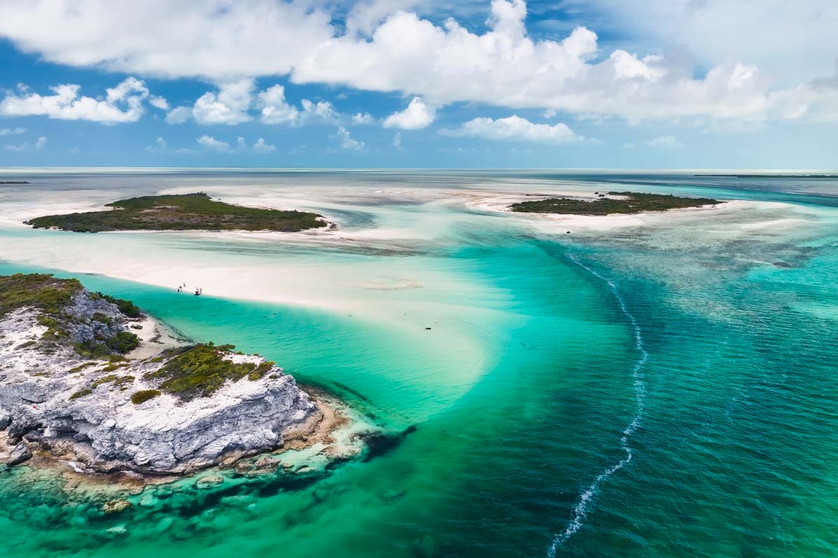 Aerial view of a tropical coastal landscape with turquoise water, sandy beaches, rocky outcrops, and green islets under a partly cloudy sky.