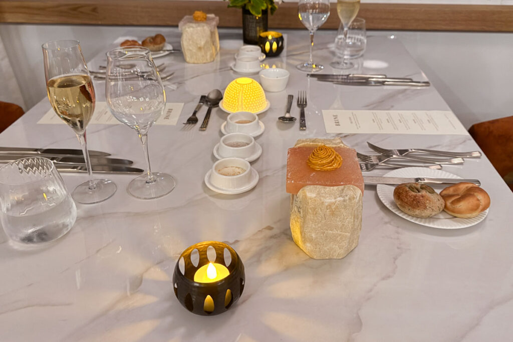 Elegant restaurant table set with glassware, utensils, bread rolls, small dishes, menus, and lit candles, featuring a decorative butter swirl on a stone slab.