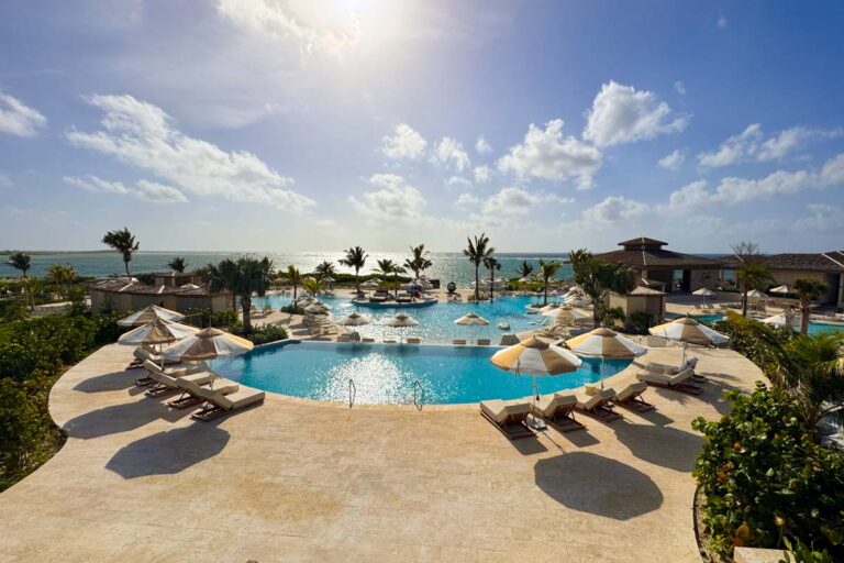 Infinity pool with sun loungers and umbrellas overlooks the ocean, surrounded by palm trees and resort buildings under a sunny, partly cloudy sky.