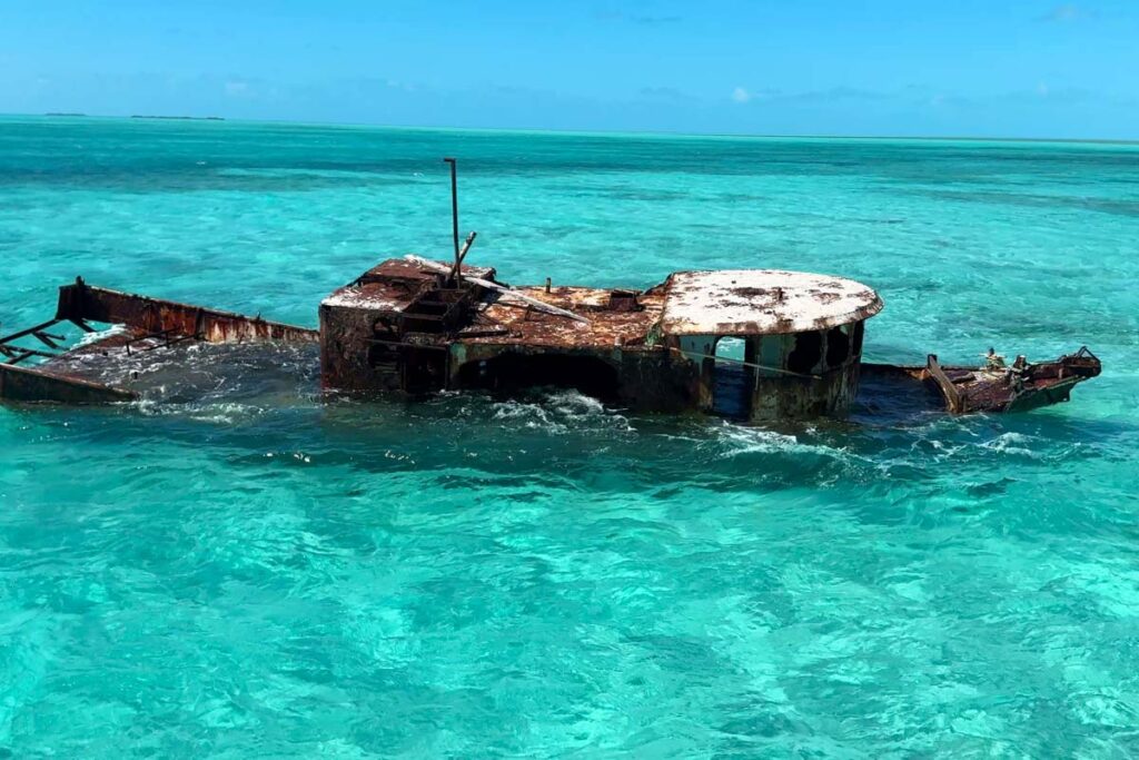 A rusted shipwreck partially submerged in clear, shallow turquoise water under a bright blue sky.