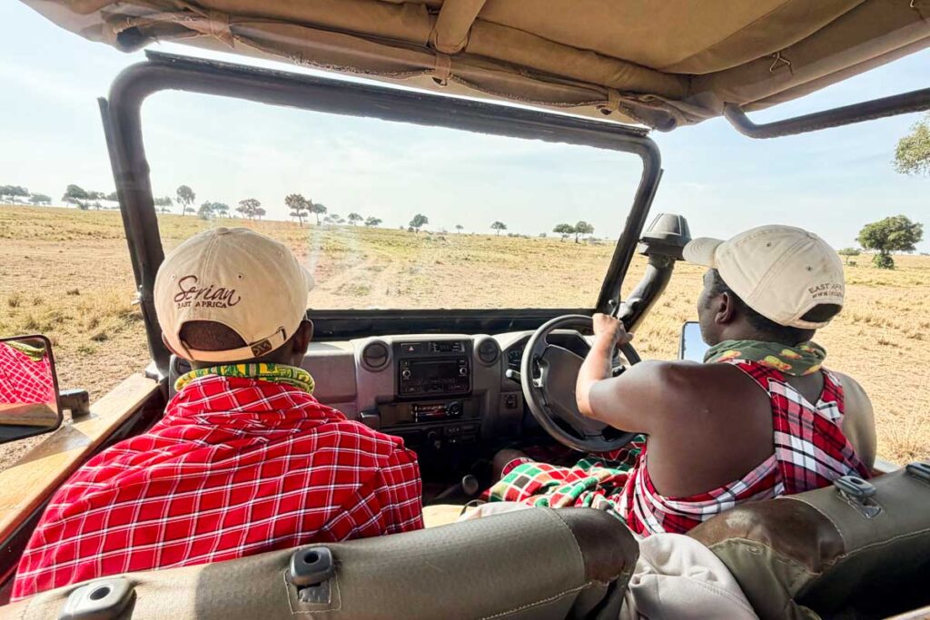 Two people wearing hats and patterned clothing sit in a safari vehicle, driving through a dry, open landscape with scattered trees.