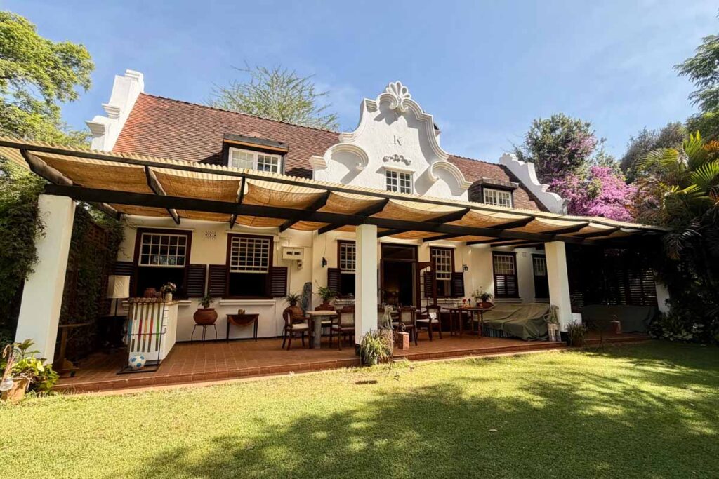 Large white house with Dutch gable roof, covered patio area with tables and chairs, set in a green garden with trees and plants.